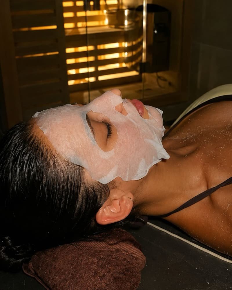 Mujer relajándose con una mascarilla facial, tumbada sobre mármol negro, con la sauna al fondo
